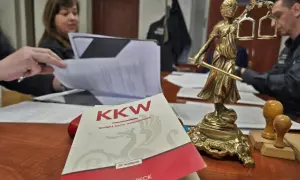 Na biurku leży Kodeks karny wykonawczy, obok Statua Temidy. W tle widać osoby pracujące nad dokumentami, co może sugerować sprawy związane z dozorem elektronicznym.