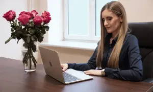 Kobieta w czarnej koszuli pracuje na laptopie przy biurku, obok bukiet róż w wazonie.