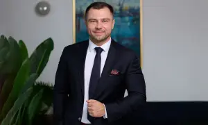 Mężczyzna w garniturze, stojący obok rośliny, z obrazem portowym w tle.