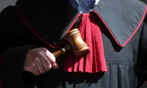 Sędzia trzyma młotek, gotowy do ogłoszenia wyroku. W tle widać togę z czerwonym żabotem, symbolizującą sprawiedliwość i powagę, która towarzyszy wydaniu subsydiarnego aktu oskarżenia.