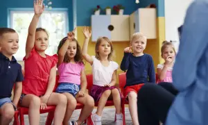 Dzieci na czerwonych krzesłach podnoszą ręce, jakby odpowiadały na pytanie, czy zerówka jest obowiązkowa.