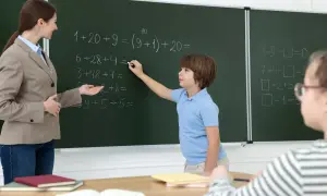 Nauczycielka pomaga uczniowi rozwiązać zadania matematyczne przy tablicy. Dzieci uczą się, wypełniając swój obowiązek szkolny.