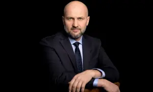 Łysy mężczyzna w garniturze i krawacie, oparty o krzesło, na czarnym tle.
