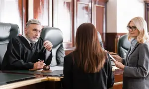 Sędzia w todze słucha zeznań, gdy prawniczka notuje. Rozprawa sądowa w toku.