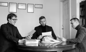 Trzech mężczyzn przy okrągłym stole analizuje dokumenty. Jeden z nich trzyma kopertę, jakby właśnie otrzymał wyrok w zawieszeniu.