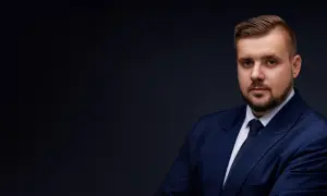 Mężczyzna w granatowym garniturze i krawacie, z zarostem, na ciemnym tle.