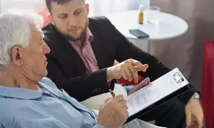Starszy mężczyzna w piżamie podpisuje dokument. Obok siedzi młodszy mężczyzna, wskazując palcem na tekst. To może być zapis windykacyjny.