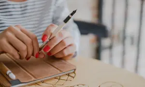 Dłonie z czerwonymi paznokciami trzymają pióro nad notatnikiem. Idealny moment, by zacząć zastanawiać się, jak napisać recenzję książki.