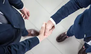 Dwóch mężczyzn w garniturach ściska sobie dłonie na jasnym tle.