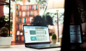 Laptop z danymi finansowymi, w tym wykresem i tabelami, prezentuje excel indeks. Obok stoi tablet, a w tle widać regały z książkami.
