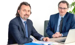 Dwóch mężczyzn w garniturach siedzi przy biurku z dokumentami i laptopem.