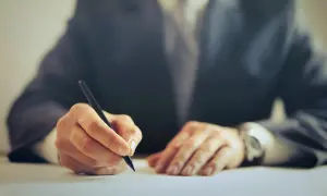 Dłoń w garniturze pisząca długopisem na białym papierze.