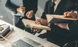 Trzy osoby w garniturach analizują dokumenty i książki przy biurku z laptopem i wagą sprawiedliwości.