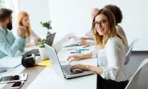 Uśmiechnięta pracownica samorządowa w okularach pracuje przy laptopie w nowoczesnym biurze.