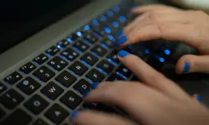 Dłonie z niebieskim lakierem na paznokciach piszą na podświetlanej klawiaturze MacBooka. Widać klawisze "twarda spacja mac".