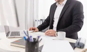 Mężczyzna w garniturze pracuje przy komputerze. Na biurku laptop, myszka, klawiatura, kubek kawy i przybory piśmiennicze. Może to być przygotowanie do wydania wyroku zaocznego.