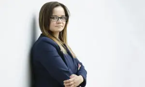 Kobieta w okularach i granatowej marynarce stoi oparta o ścianę, z rękami skrzyżowanymi na piersi.