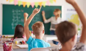 Uczniowie z podniesionymi rękami, gotowi do odpowiedzi. Wychowawca stoi przy tablicy, tłumacząc lekcję.