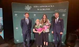 Cztery osoby w garniturach i sukience stoją na scenie z bukietami kwiatów przed zielonym banerem.