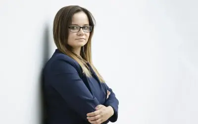 Kobieta w okularach i granatowej marynarce stoi oparta o ścianę, z rękami skrzyżowanymi na piersi.