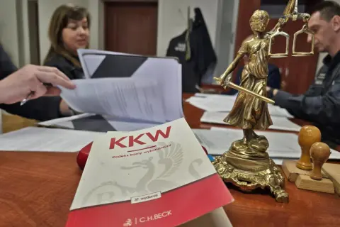 Na biurku leży Kodeks karny wykonawczy, obok Statua Temidy. W tle widać osoby pracujące nad dokumentami, co może sugerować sprawy związane z dozorem elektronicznym.