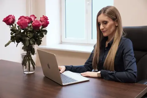 Kobieta w czarnej koszuli pracuje na laptopie przy biurku, obok bukiet róż w wazonie.