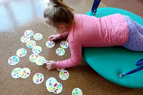 Dziewczynka na huśtawce bawi się kartami z cyframi i kształtami. To element jej rewalidacji, wspierający rozwój.