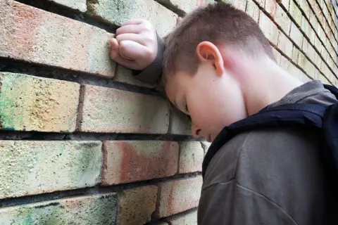 Chłopiec z plecakiem opiera głowę o ceglaną ścianę, wyrażając smutek i poczucie osamotnienia, które mogą być objawami fobii szkolnej.
