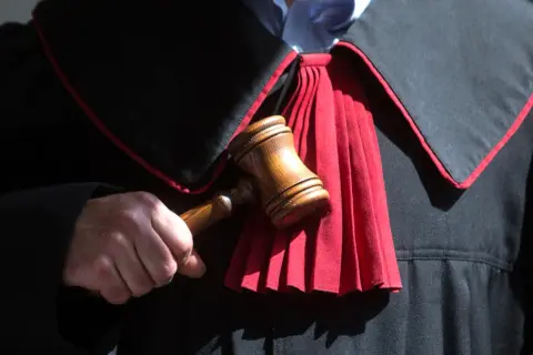 Sędzia trzyma młotek, gotowy do ogłoszenia wyroku. W tle widać togę z czerwonym żabotem, symbolizującą sprawiedliwość i powagę, która towarzyszy wydaniu subsydiarnego aktu oskarżenia.