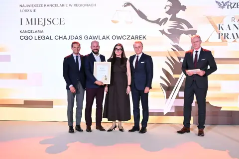 Pięć osób w strojach formalnych pozuje do zdjęcia na tle sceny z grafiką i napisem "I MIEJSCE".