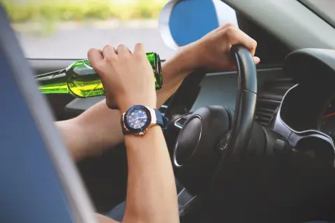 Ręce na kierownicy, jedna trzyma butelkę piwa. Ryzykowne zachowanie, stan po użyciu alkoholu.