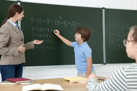 Nauczycielka pomaga uczniowi rozwiązać zadania matematyczne przy tablicy. Dzieci uczą się, wypełniając swój obowiązek szkolny.