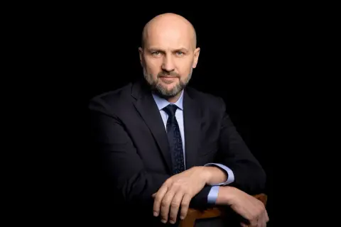 Łysy mężczyzna w garniturze i krawacie, oparty o krzesło, na czarnym tle.