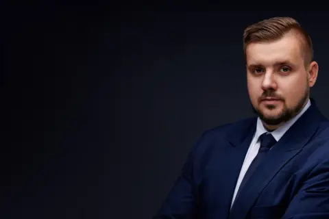 Mężczyzna w granatowym garniturze i krawacie, z zarostem, na ciemnym tle.