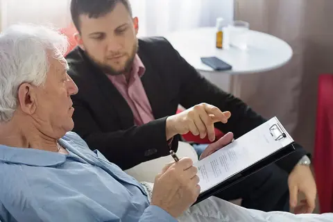 Starszy mężczyzna w piżamie podpisuje dokument. Obok siedzi młodszy mężczyzna, wskazując palcem na tekst. To może być zapis windykacyjny.