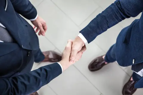 Dwóch mężczyzn w garniturach ściska sobie dłonie na jasnym tle.