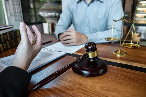 Młotek sędziowski, waga sprawiedliwości i ręka trzymająca długopis na tle dokumentów.