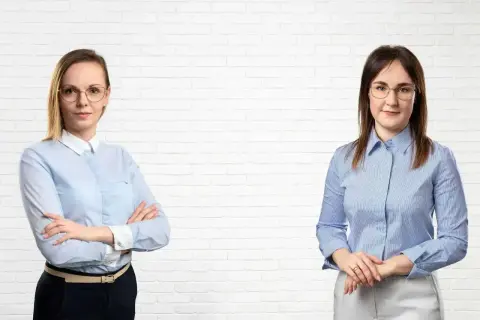 Dwie kobiety w koszulach i okularach na tle białej ceglanej ściany.