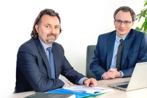 Dwóch mężczyzn w garniturach siedzi przy biurku z dokumentami i laptopem.