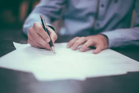 Dłoń pisze piórem na dokumencie, być może to dobrowolne poddanie się karze.