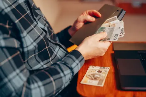 Osoba w kratę liczy pieniądze, przygotowując donos do urzędu skarbowego. Obok leży laptop.