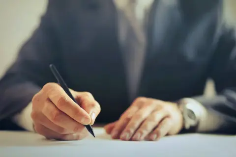 Dłoń w garniturze pisząca długopisem na białym papierze.