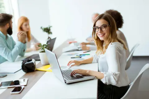 Uśmiechnięta pracownica samorządowa w okularach pracuje przy laptopie w nowoczesnym biurze.
