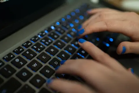 Dłonie z niebieskim lakierem na paznokciach piszą na podświetlanej klawiaturze MacBooka. Widać klawisze "twarda spacja mac".
