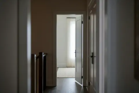 Korytarz z otwartymi drzwiami, sugerującymi naruszenie miru domowego. W tle jasne zasłony i dywan.