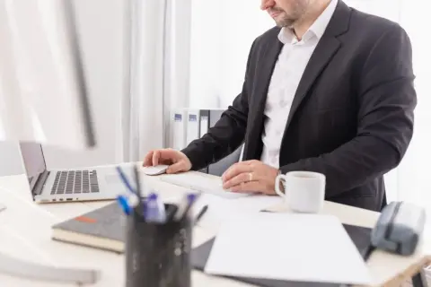 Mężczyzna w garniturze pracuje przy komputerze. Na biurku laptop, myszka, klawiatura, kubek kawy i przybory piśmiennicze. Może to być przygotowanie do wydania wyroku zaocznego.