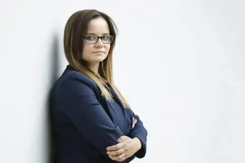 Kobieta w okularach i granatowej marynarce stoi oparta o ścianę, z rękami skrzyżowanymi na piersi.