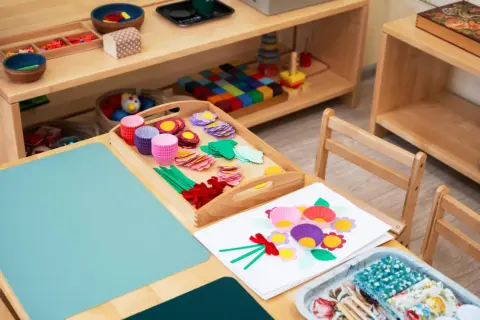 Zestaw do tworzenia kwiatów metodą Montessori: kolorowe papierowe foremki, listki, kółka i wstążki na drewnianej tacy.