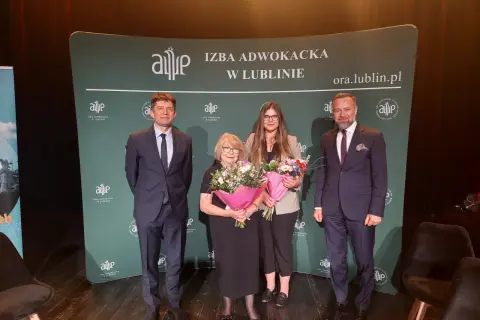Cztery osoby w garniturach i sukience stoją na scenie z bukietami kwiatów przed zielonym banerem.