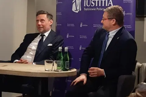 Dwóch mężczyzn w garniturach siedzi przy stole, za nimi fioletowe tło z logo "JUSTITIA".
